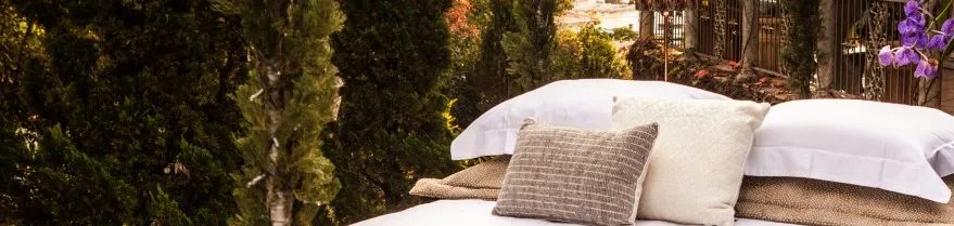 Cama com roupa de cama branca e almofadas em tecelagem artesanal de Joel Reis, em varanda cercada de vegetação
